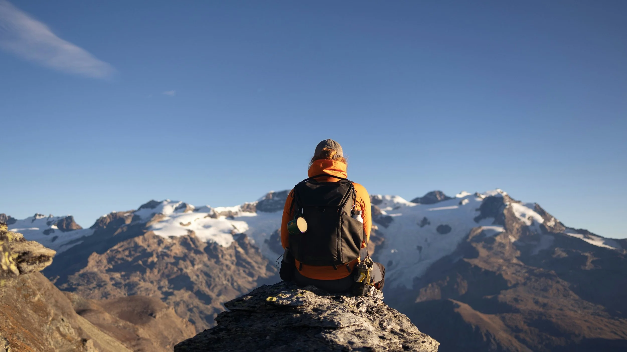 Person with a backpack sitting on a rock, overlooking snow-capped mountains under a clear blue sky.