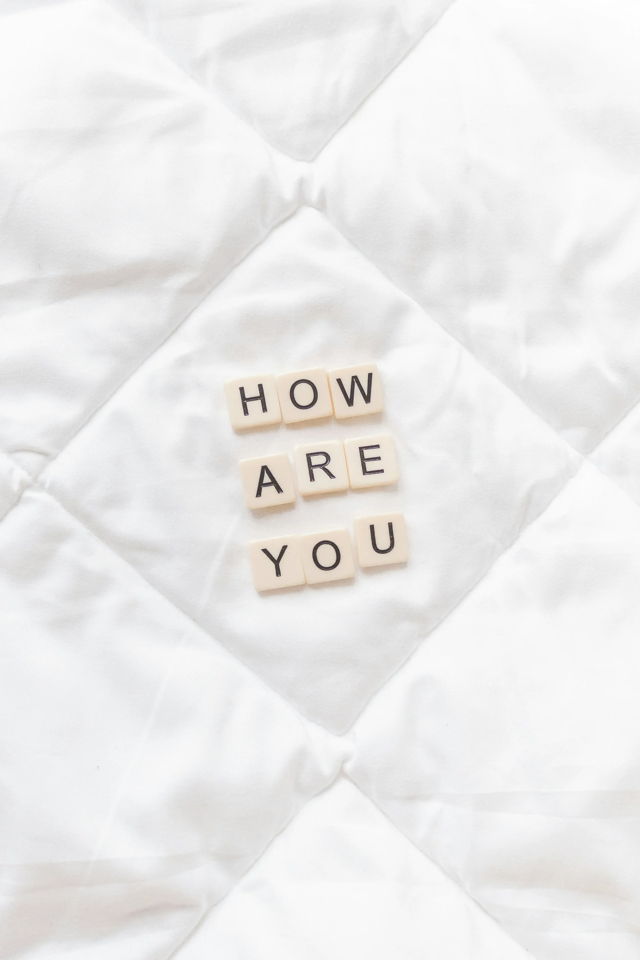 Scrabble tiles spelling out "HOW ARE YOU" on a white quilted fabric background.