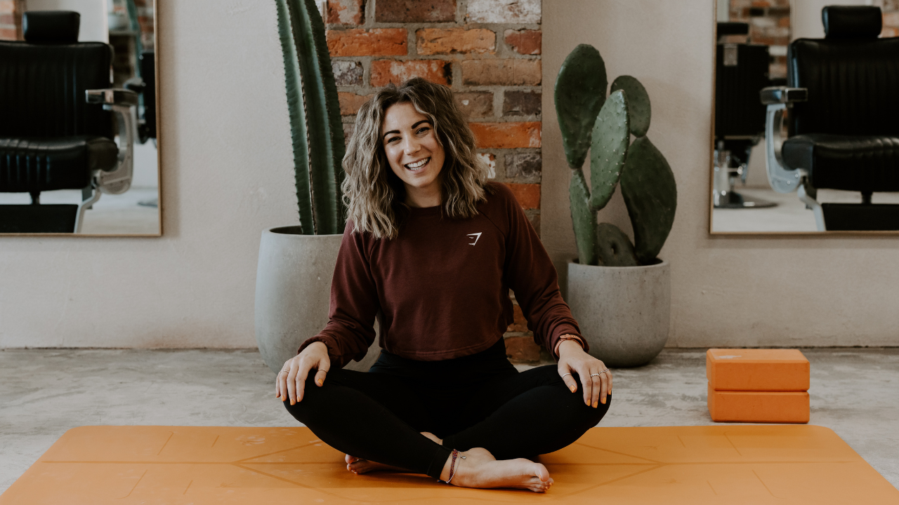 A woman with curly hair sitting cross-legged on an orange yoga mat, smiling. Behind her are two potted cacti, with a mirror reflecting black chairs and a brick wall.