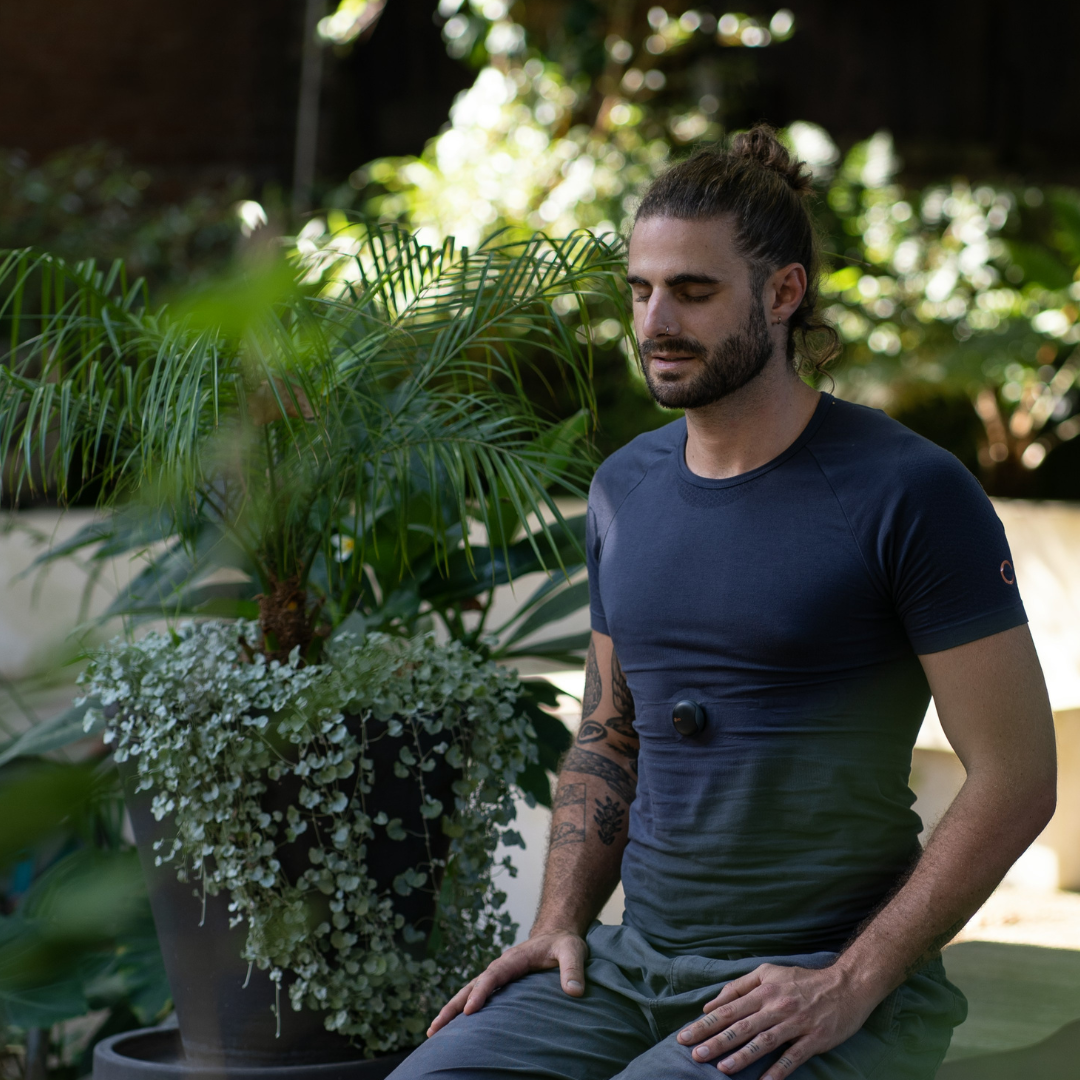 A man with long hair tied in a bun, beard, and tattoos on his left arm, sits outdoors with eyes closed, wearing a navy blue athletic shirt with a black massage ball on his chest, surrounded by green plants and sunlight filtering through trees.