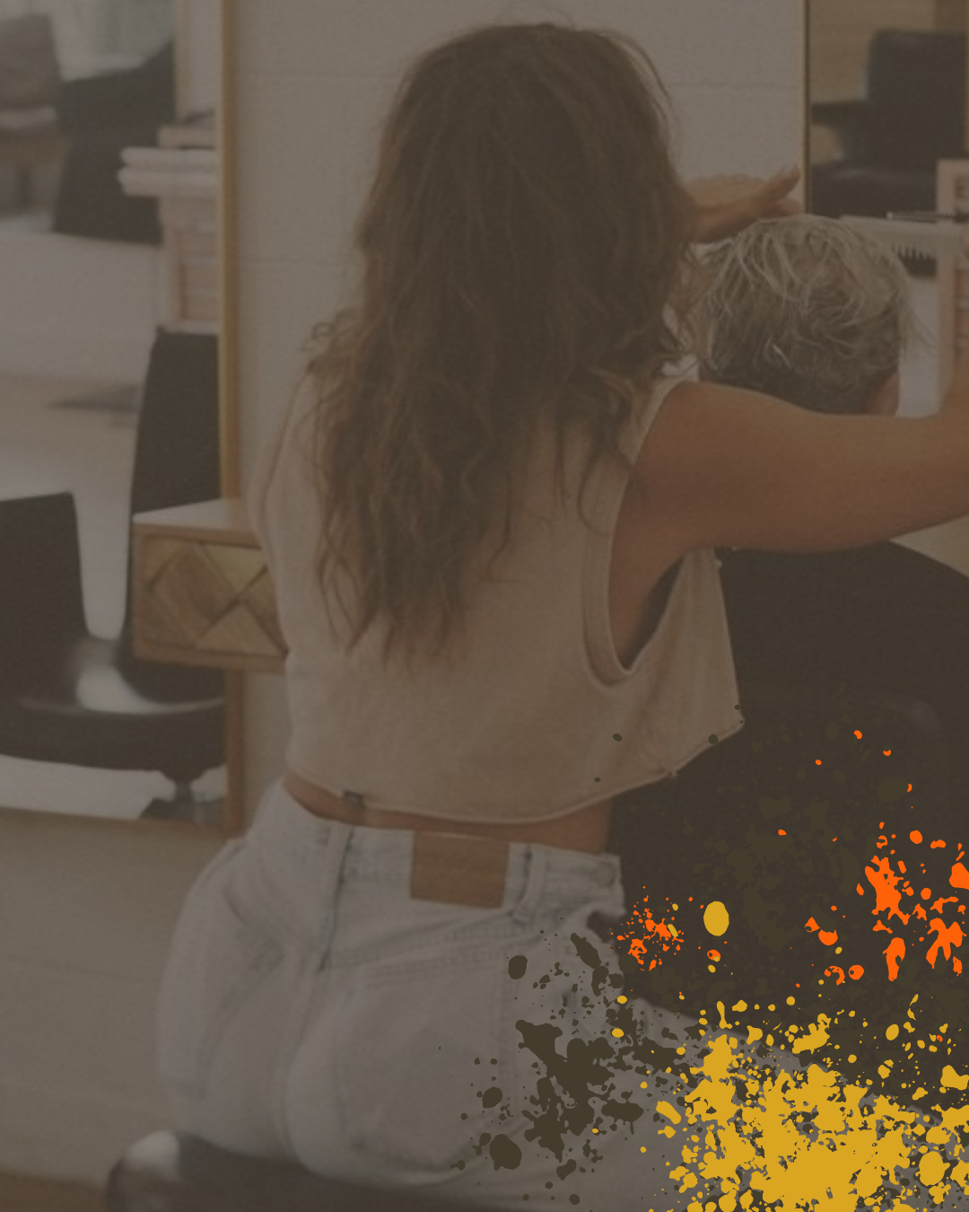 A woman with long, wavy brown hair takes a selfie in a mirror at a hair salon, wearing a sleeveless top and white jeans with a brown belt, with colorful paint splatters overlaid in the foreground.