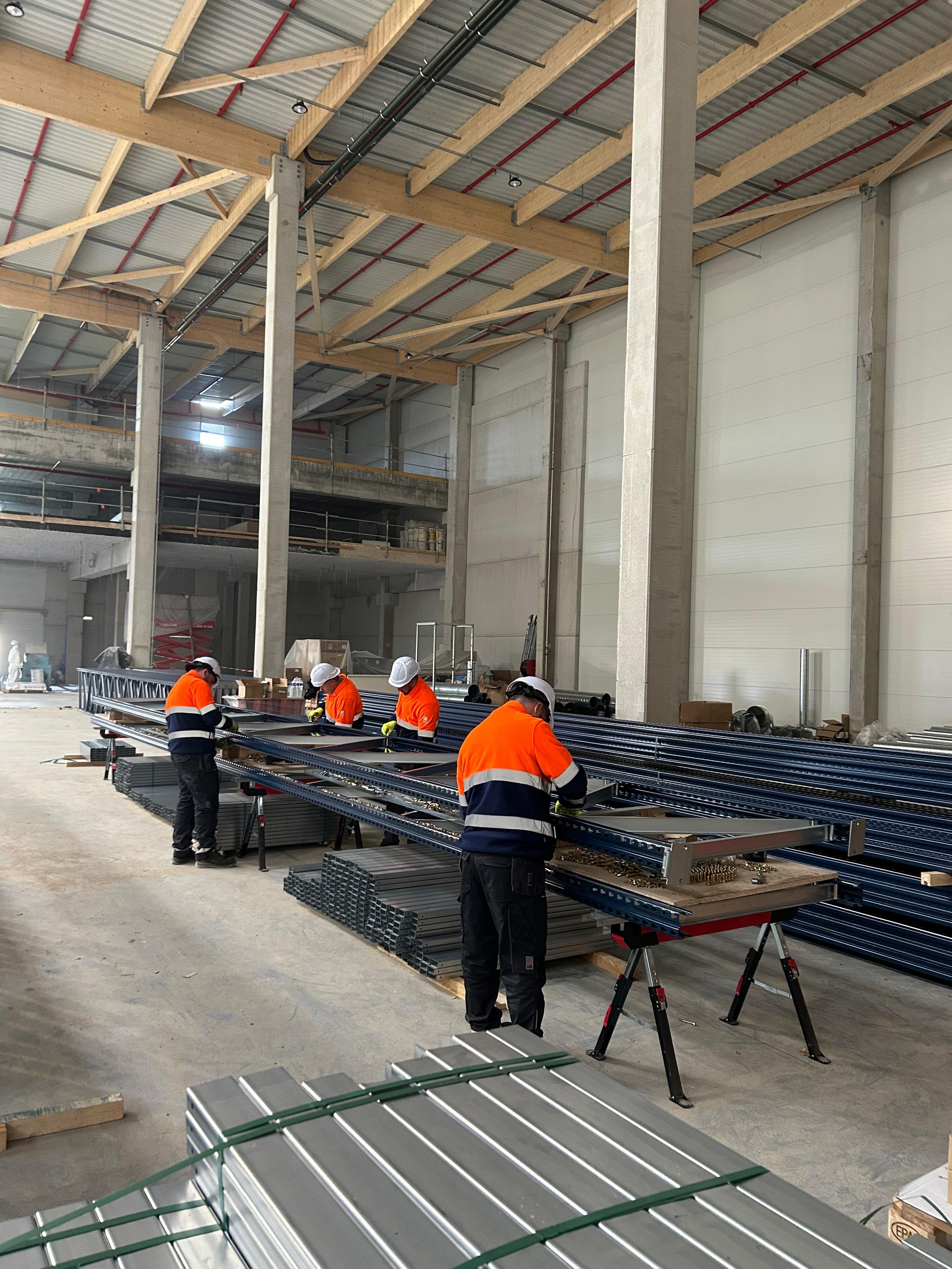 Quatre ouvriers en vêtements de sécurité orange et noire travaillent sur des structures métalliques dans une usine de construction.