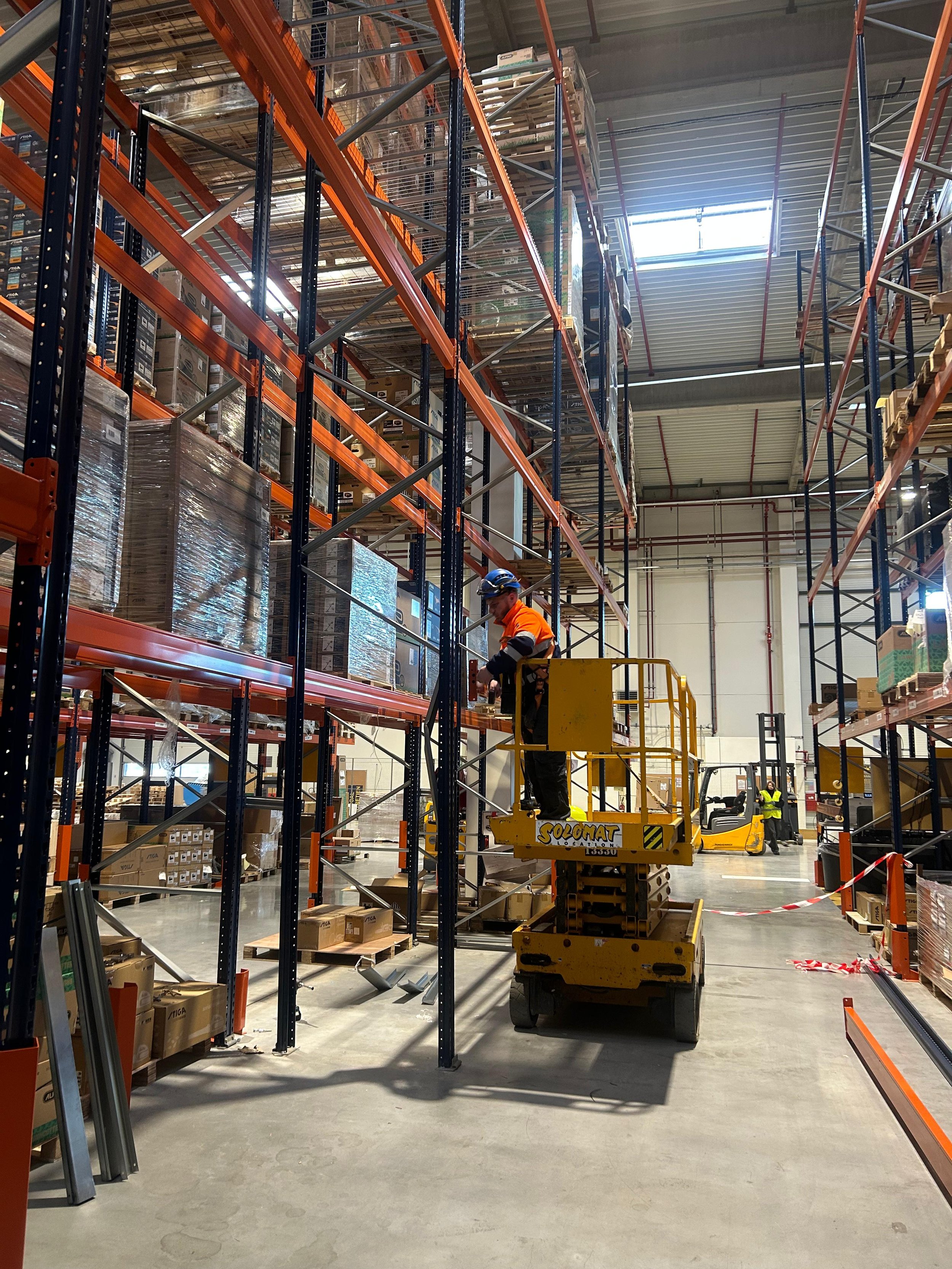 Ouvrier utilisant une plateforme élévatrice pour accéder à des rayonnages dans un grand entrepôt de stockage.