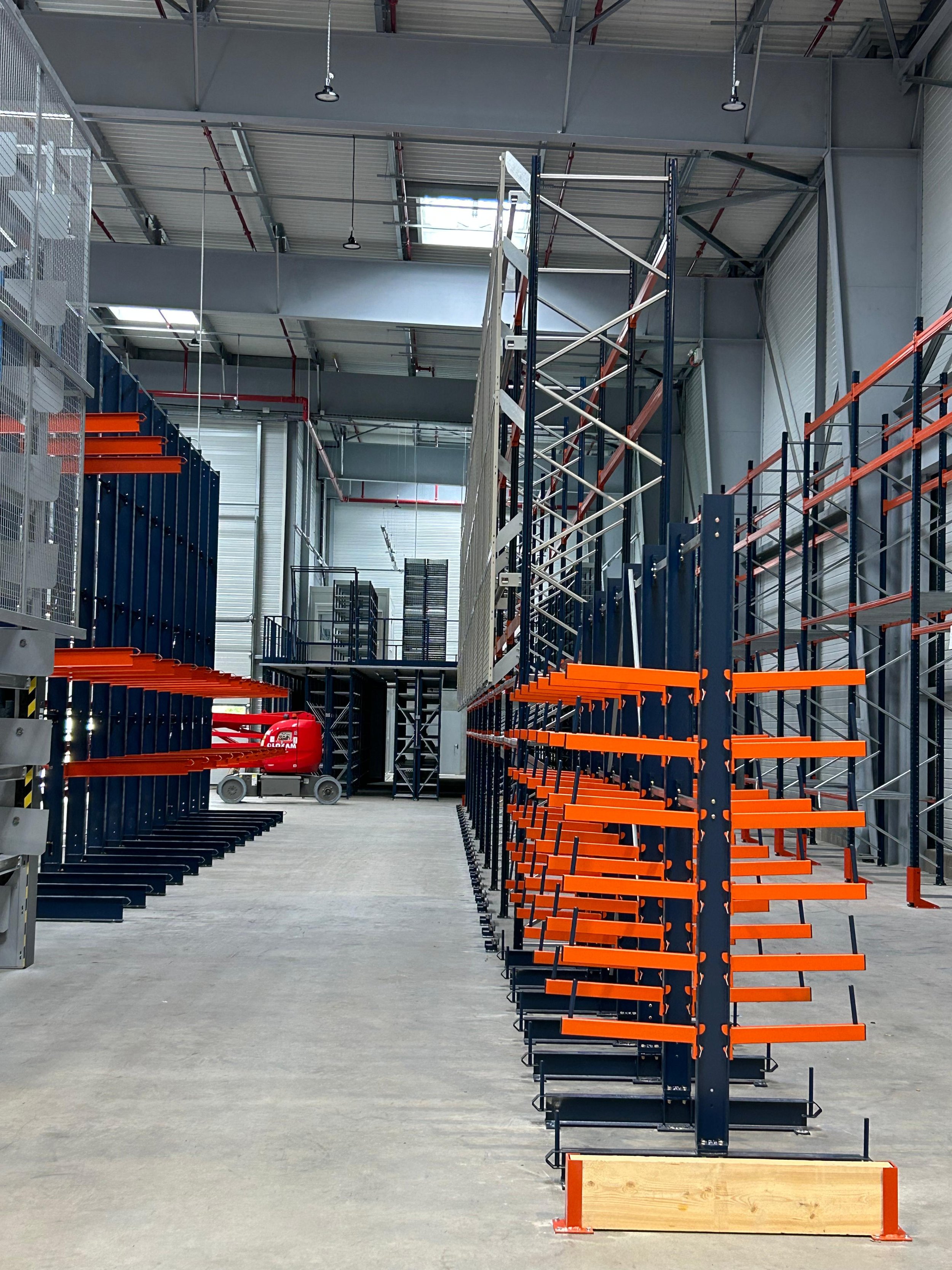 Intérieur d'un entrepôt industriel avec des étagères métalliques orange et bleues, un chariot élévateur rouge et un espace de stockage en construction.