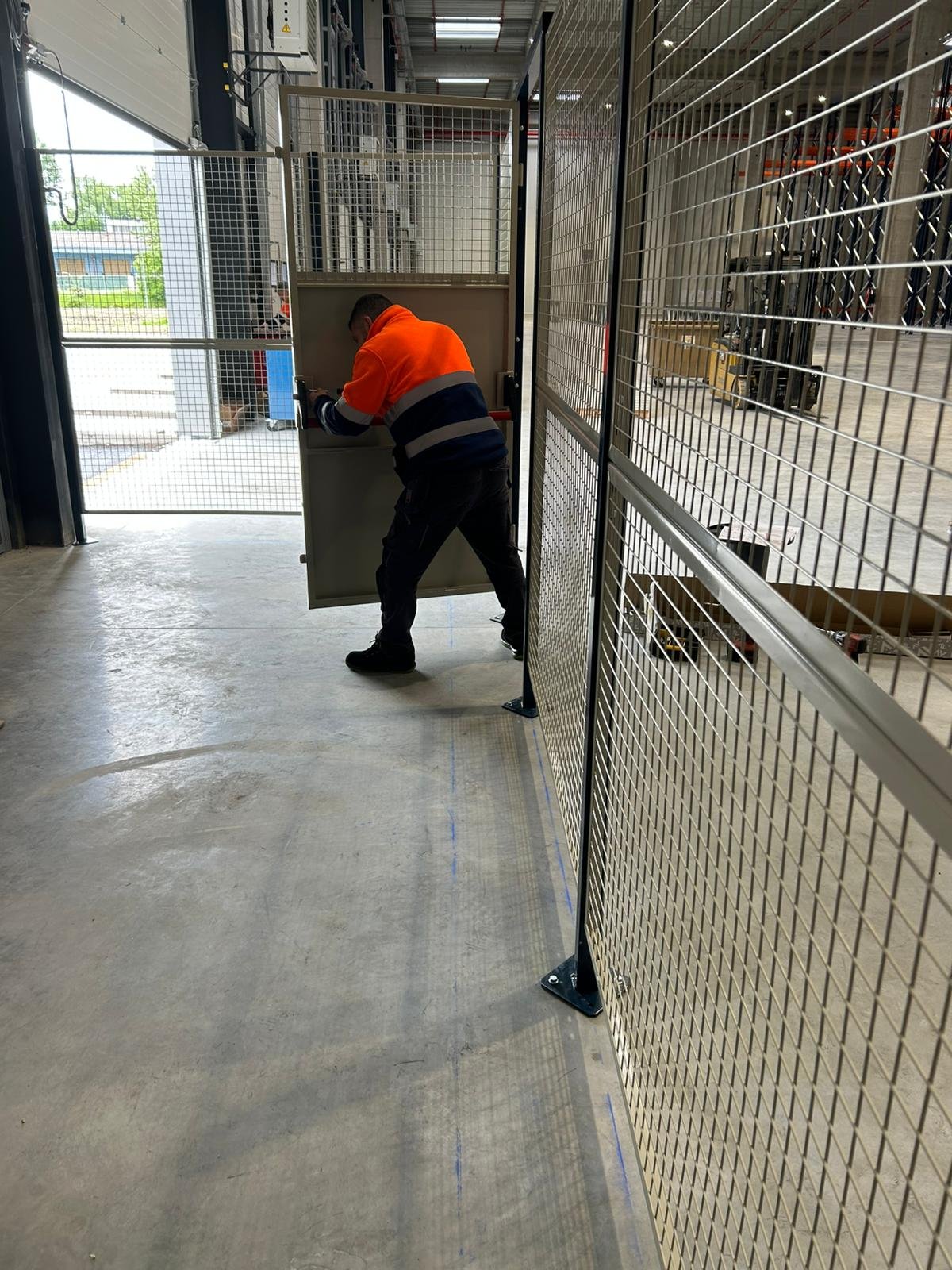 Un employé en chemise orange et pantalon noir installe une porte ou un panneau dans une zone de stockage ou d'entrepôt, entouré de grilles en métal.
