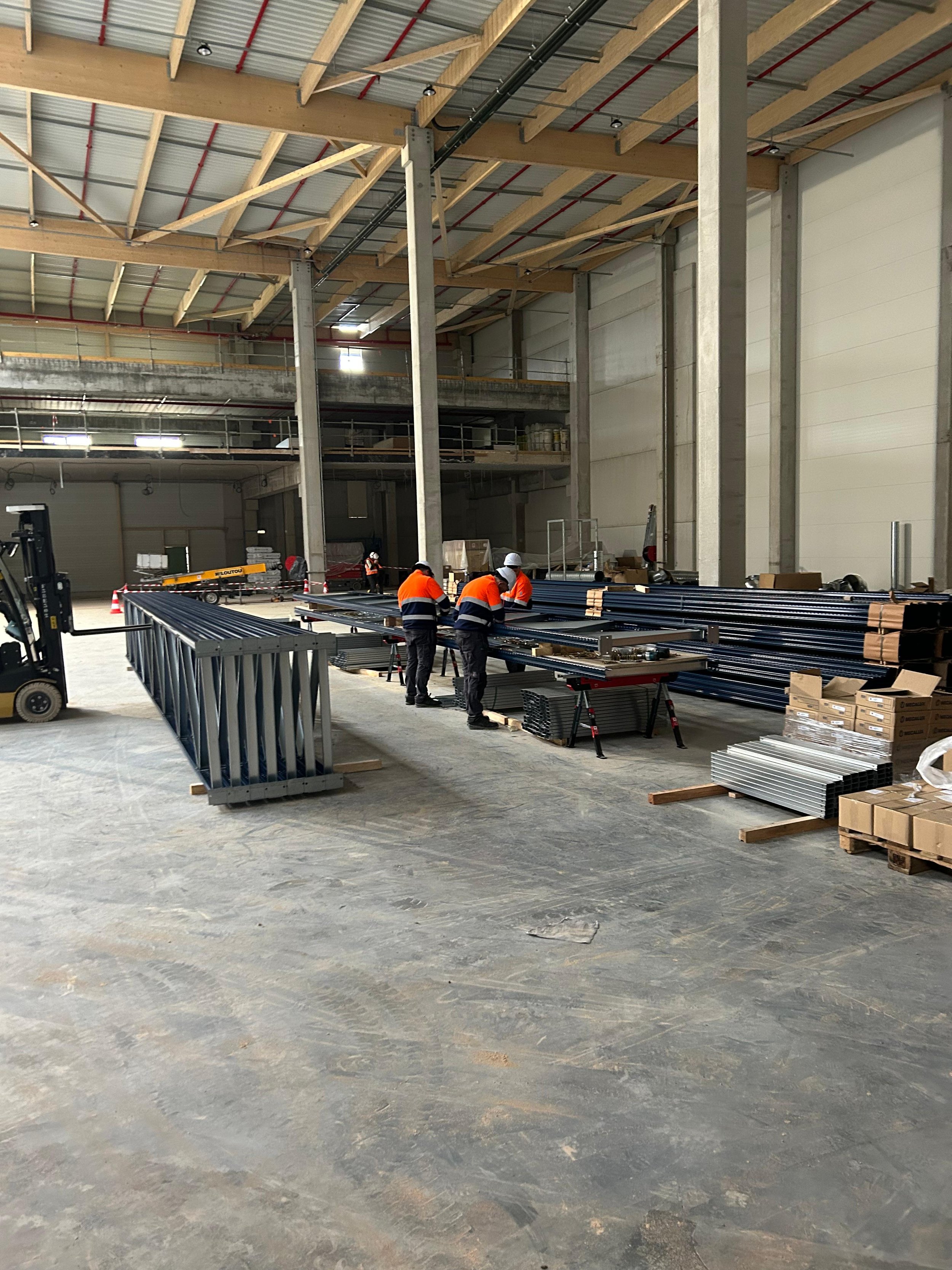 Travailleurs portant des équipements de protection travaillant à l'intérieur d'une grande structure en construction, avec du métal empilé et des matériaux de construction autour.