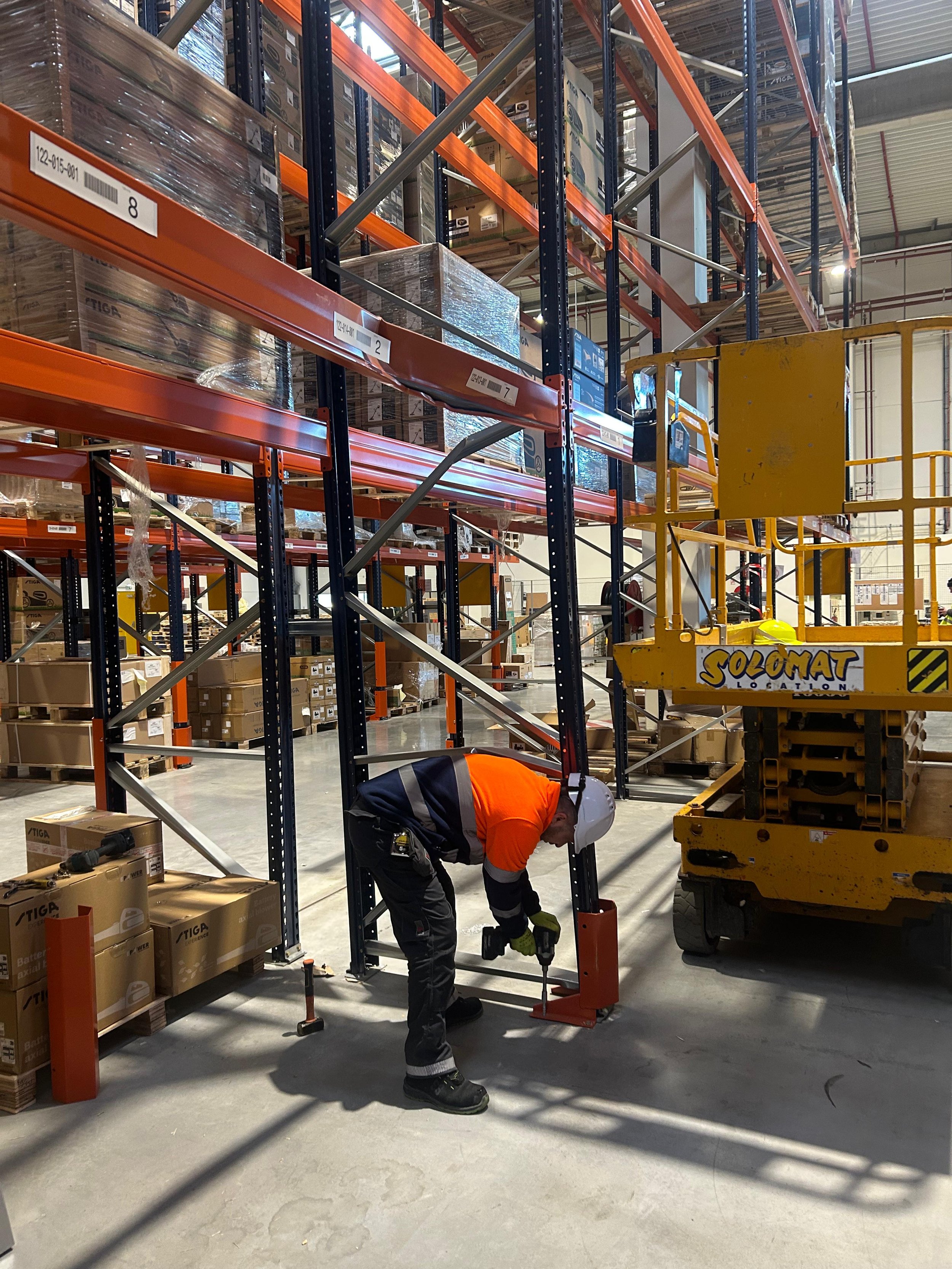 Un ouvrier en tenue de sécurité travaillant dans un entrepôt avec des étagères remplies de palettes de produits, utilisant un outil électrique, et à proximité d'une plateforme élévatrice jaune.