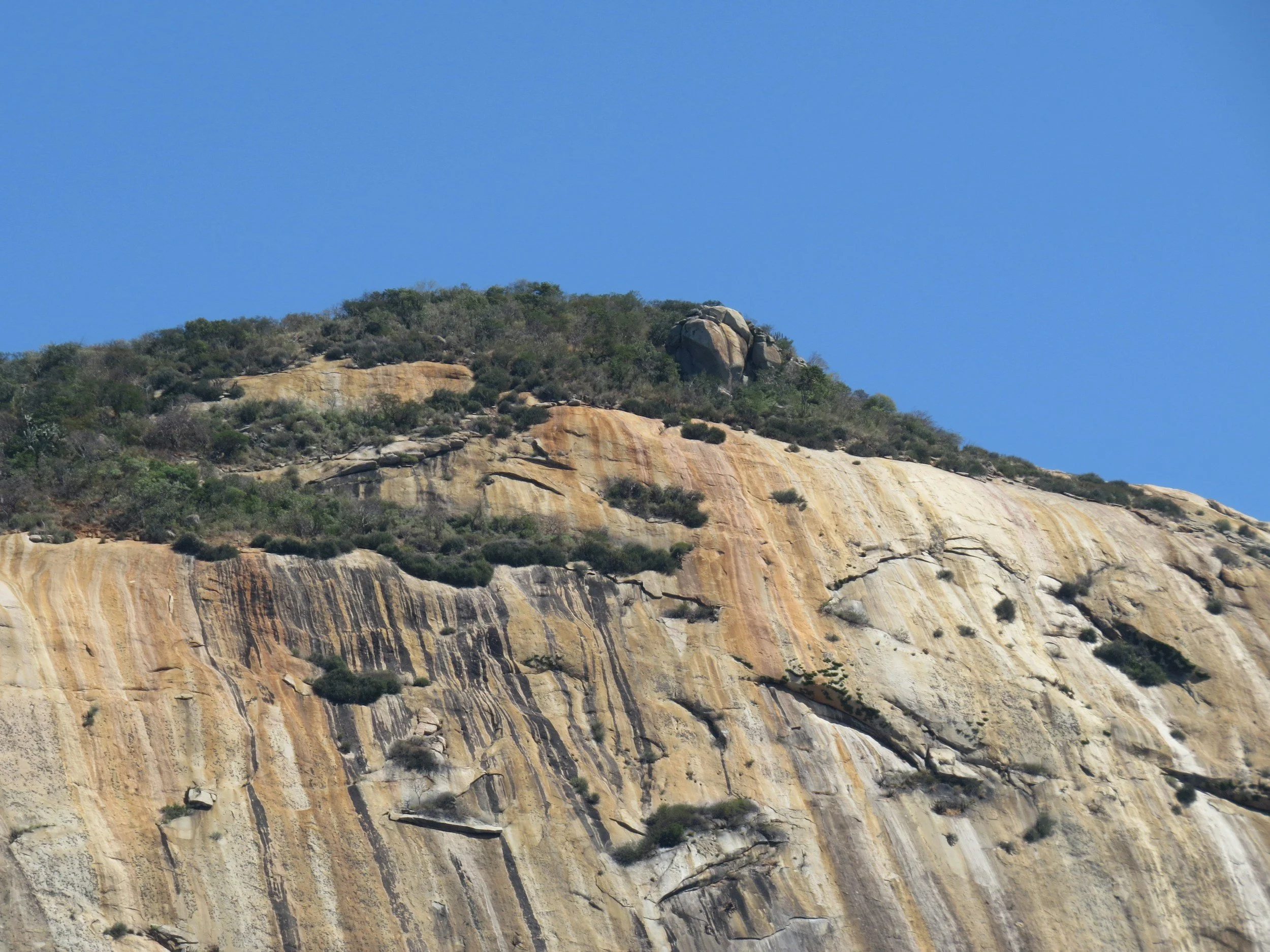 A rocky mountainside with exposed layers of sandstone and sparse vegetation against a clear blue sky.