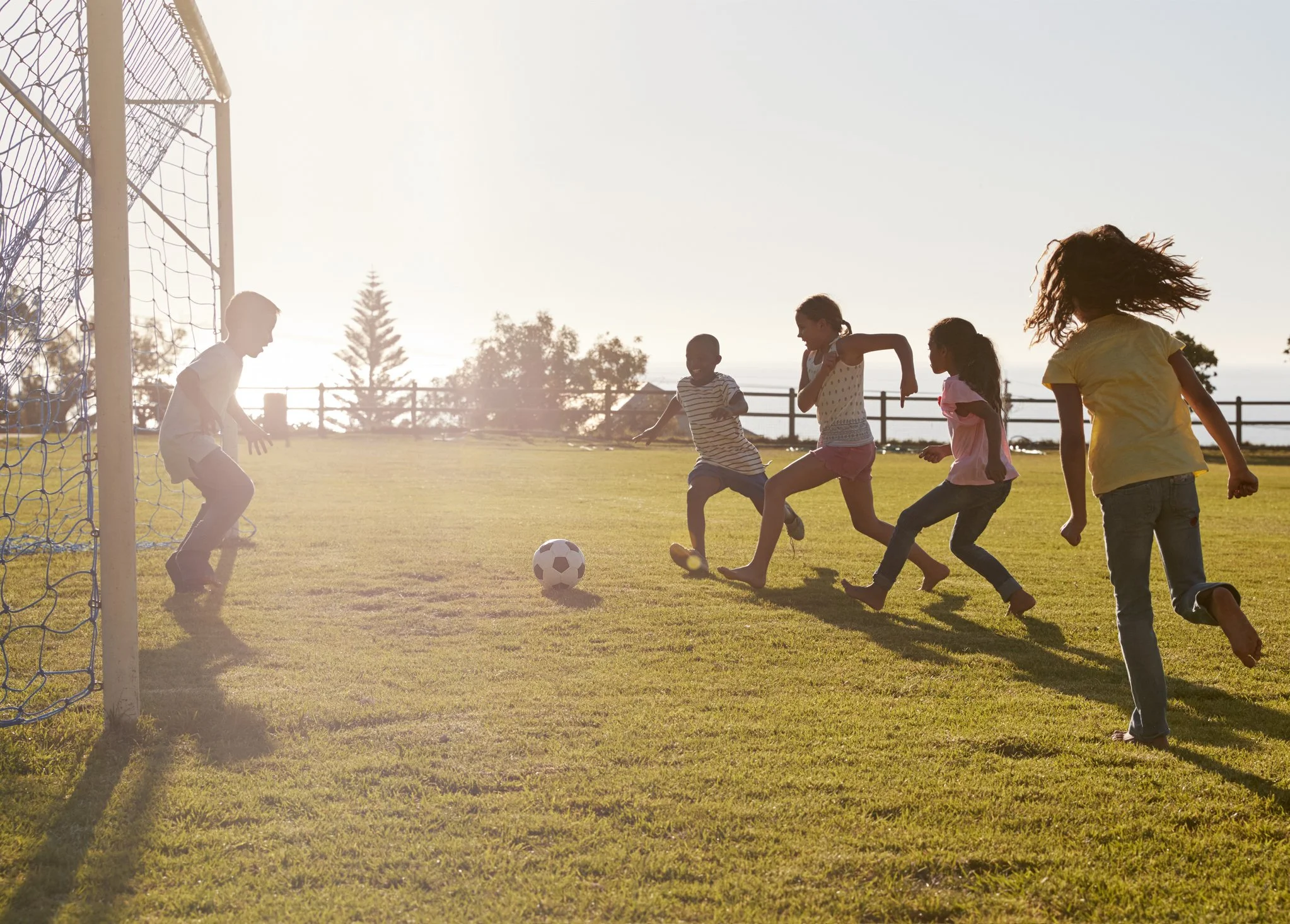 Children playing soccer on a field near a goalpost with sunlight in the background.