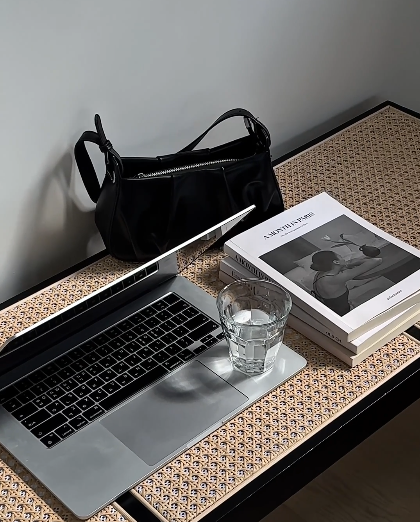 Open laptop, glass of water, stacked books, black handbag, and patterned surface on a desk.