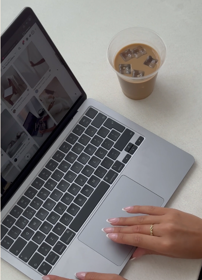 A person's hand with a gold ring on the finger, resting on a silver laptop keyboard, next to a cup of iced coffee on a white surface.