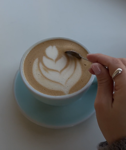 A cup of coffee with latte art in the shape of a tulip, held with a spoon and placed on a light blue saucer.