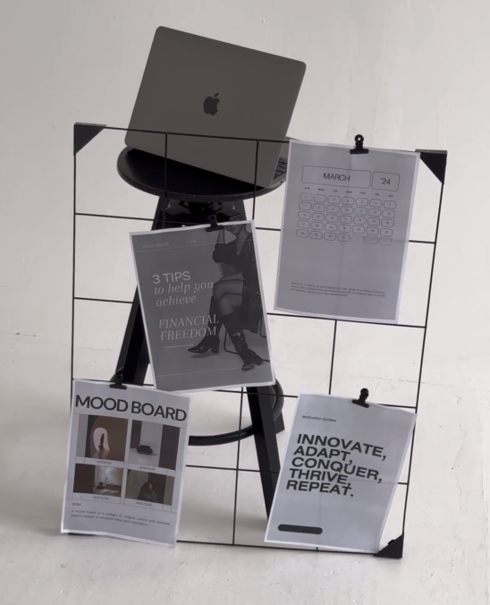 A corkboard with various printed papers pinned to it, including a calendar, an inspirational quote, a mood board, and tips for financial freedom, with a laptop placed on a stand at the top.