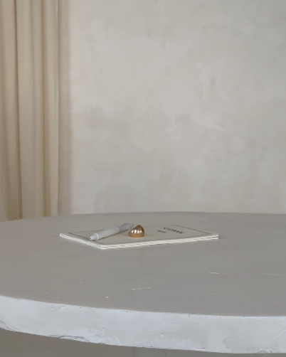 A notebook with a pen and a small decorative object on top, placed on a round gray table against a plain wall background.