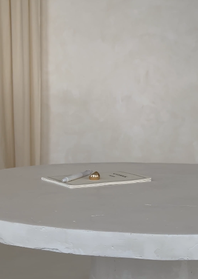 A table with a notebook, pen, and a small decorative object on top, against a plain wall background.