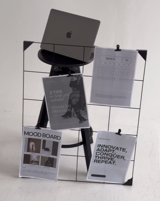 A wire grid stand with four papers attached, including a calendar page, a survey or feedback form, a motivational poster, and a mood board, with a MacBook placed on a black stand on top of the grid against a plain background.