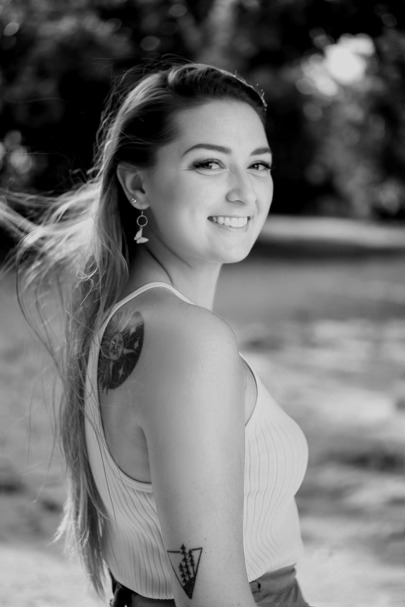 A smiling woman with long hair, wearing earrings and a sleeveless top, standing outdoors with a scenic background, in black and white.