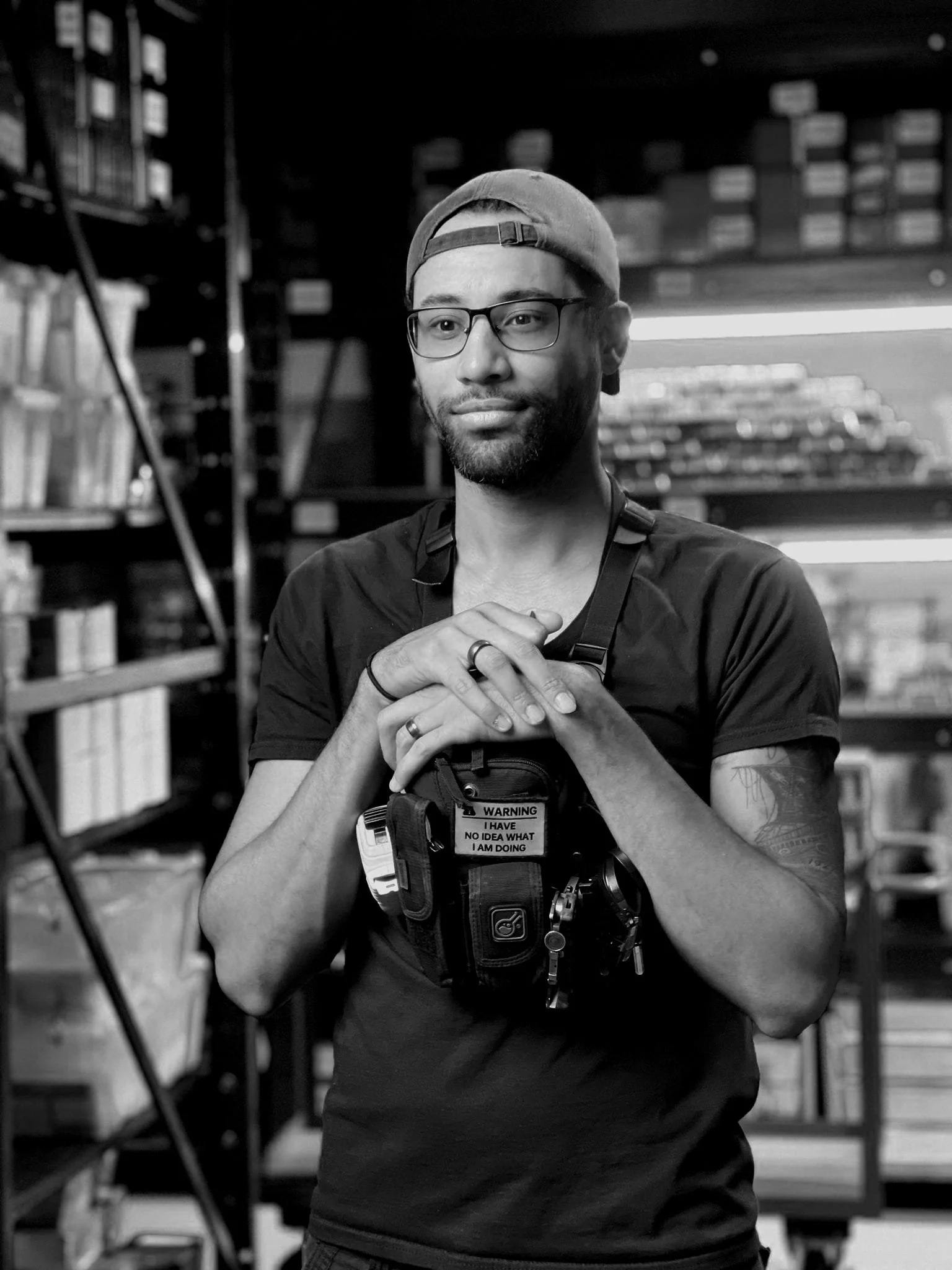 A man with glasses and a backwards cap standing in a storage room, wearing a camera or phone holder around his neck, holding his hands together in front of him.