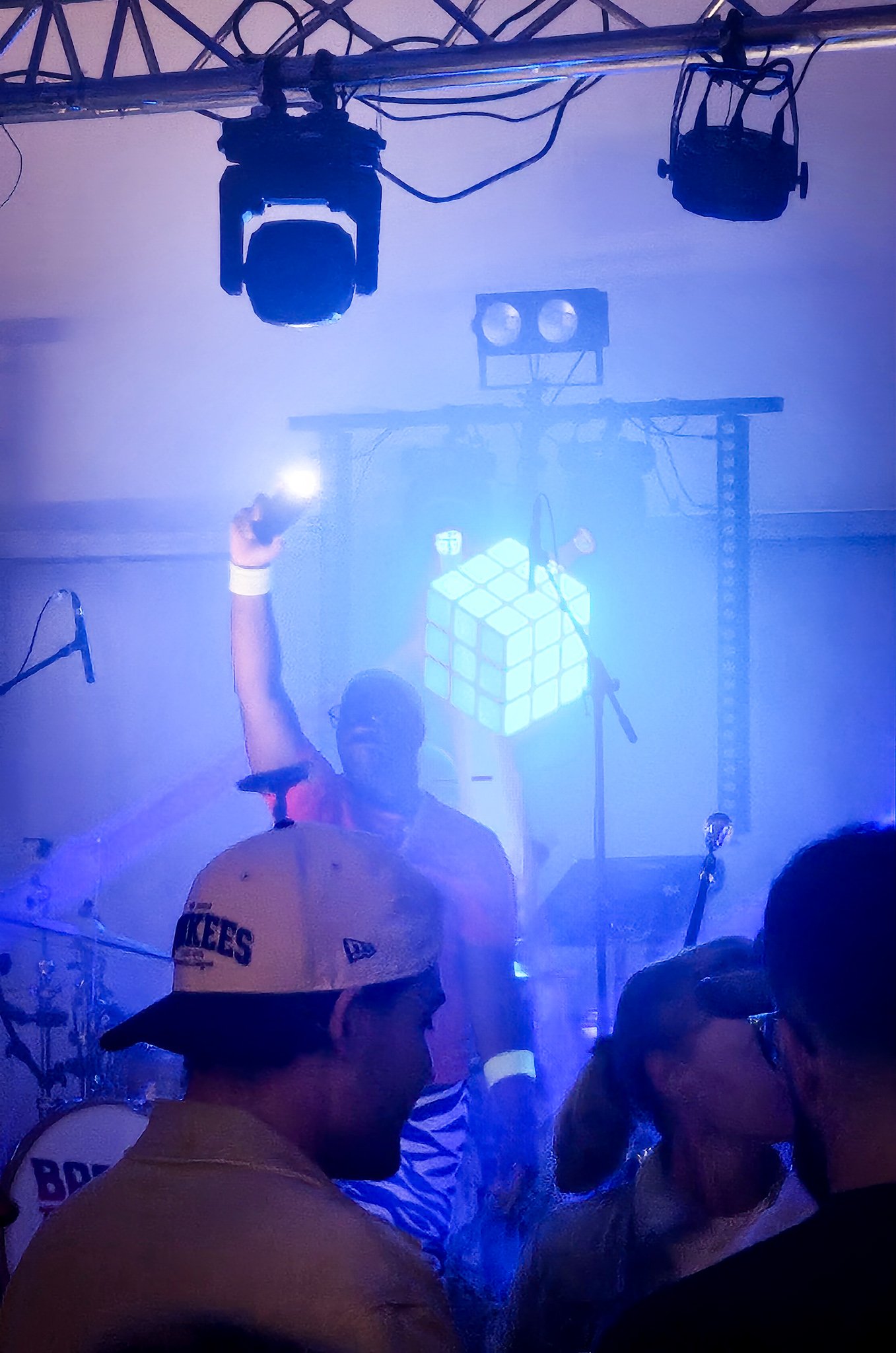 People at a concert or event with stage lighting, a person wearing a yellow baseball cap, a person in a striped shirt near a drum set, and fog or smoke effects.
