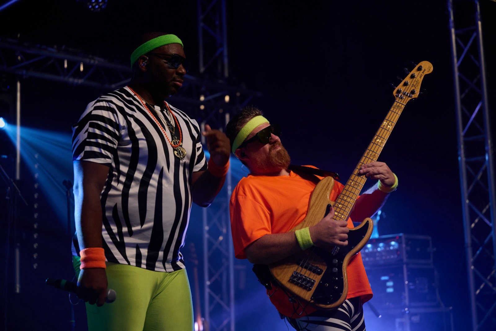 Two musicians performing on stage: a man in a zebra-patterned shirt and neon green pants holding a microphone, and a man in an orange shirt playing an electric bass guitar. Both wear neon green and black headbands and sunglasses.