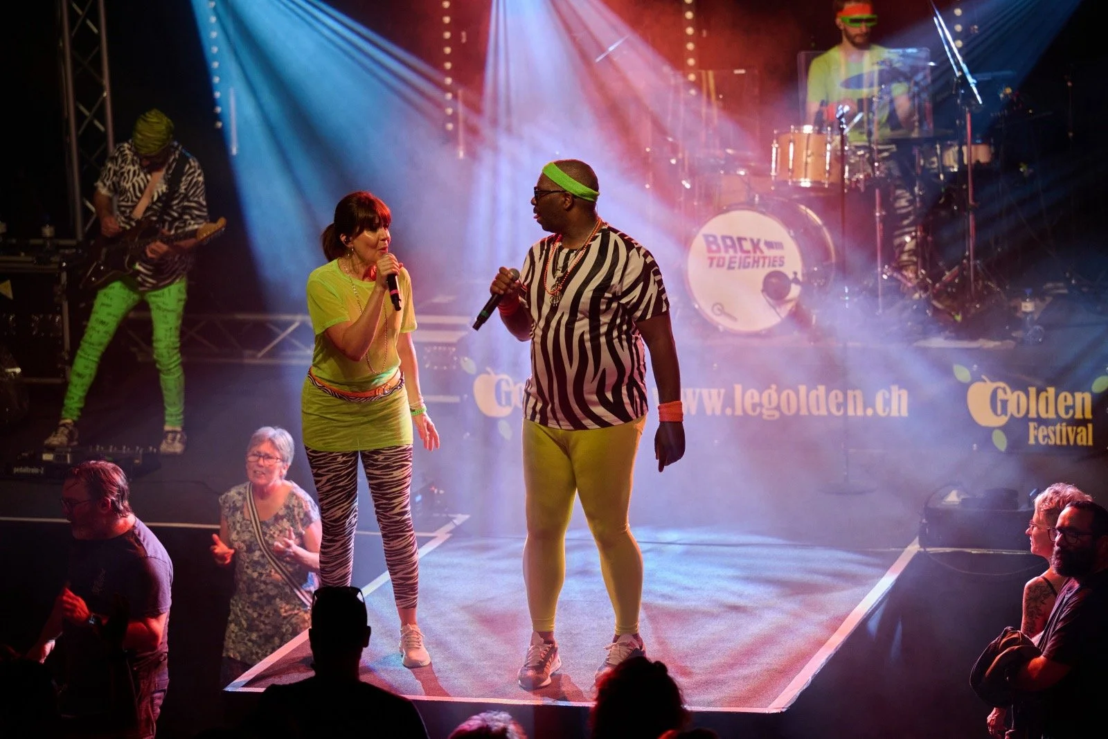 Performers on stage at the Golden Festival, dressed in zebra and animal print clothing, with colorful lighting and a live band in the background.