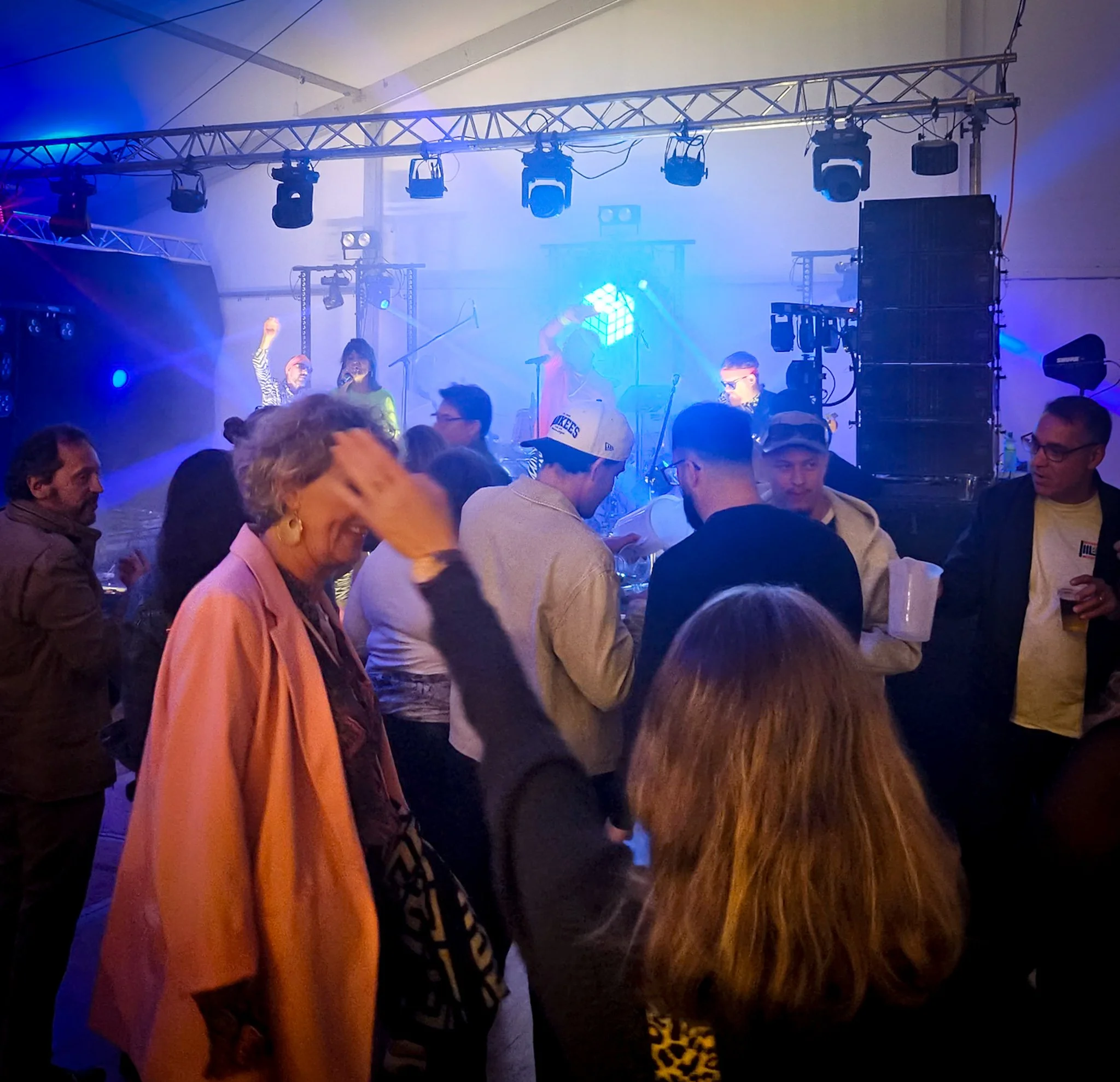 People dancing and socializing at a lively indoor concert or party with stage lighting and sound equipment.