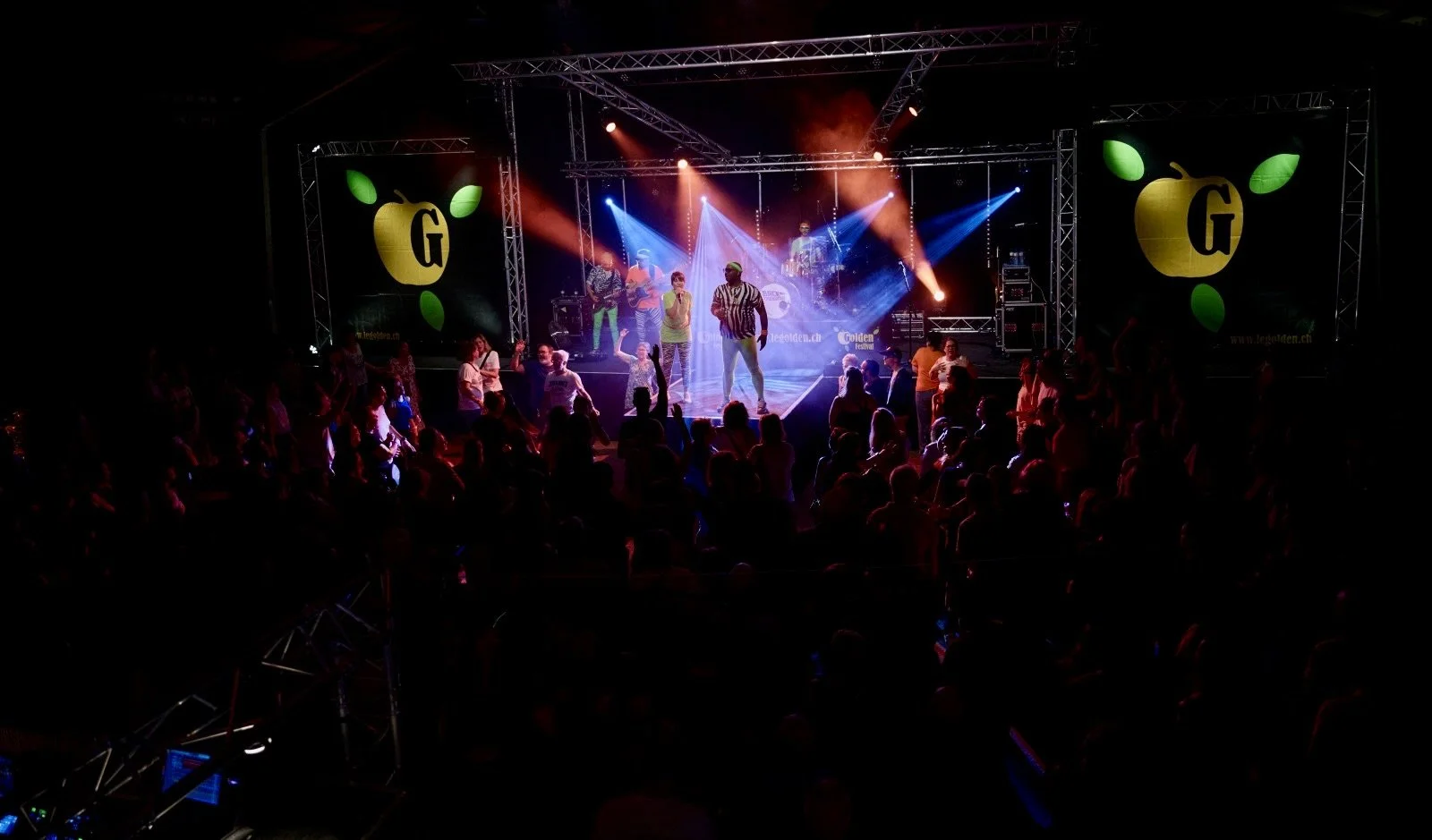 Live music concert with a band on stage, colorful lighting, audience dancing and enjoying the event, two large screens with a yellow apple logo and the letter G.