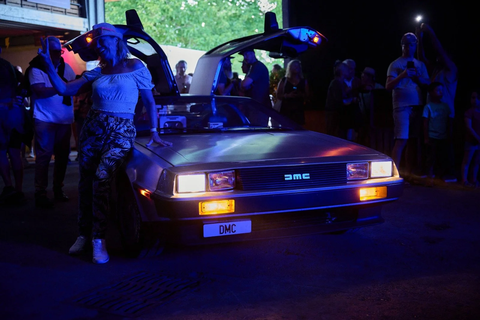 A person wearing a virtual reality headset and gesturing with one hand, standing next to a DeLorean DMC car with its gullwing door open, at a nighttime event with people in the background.