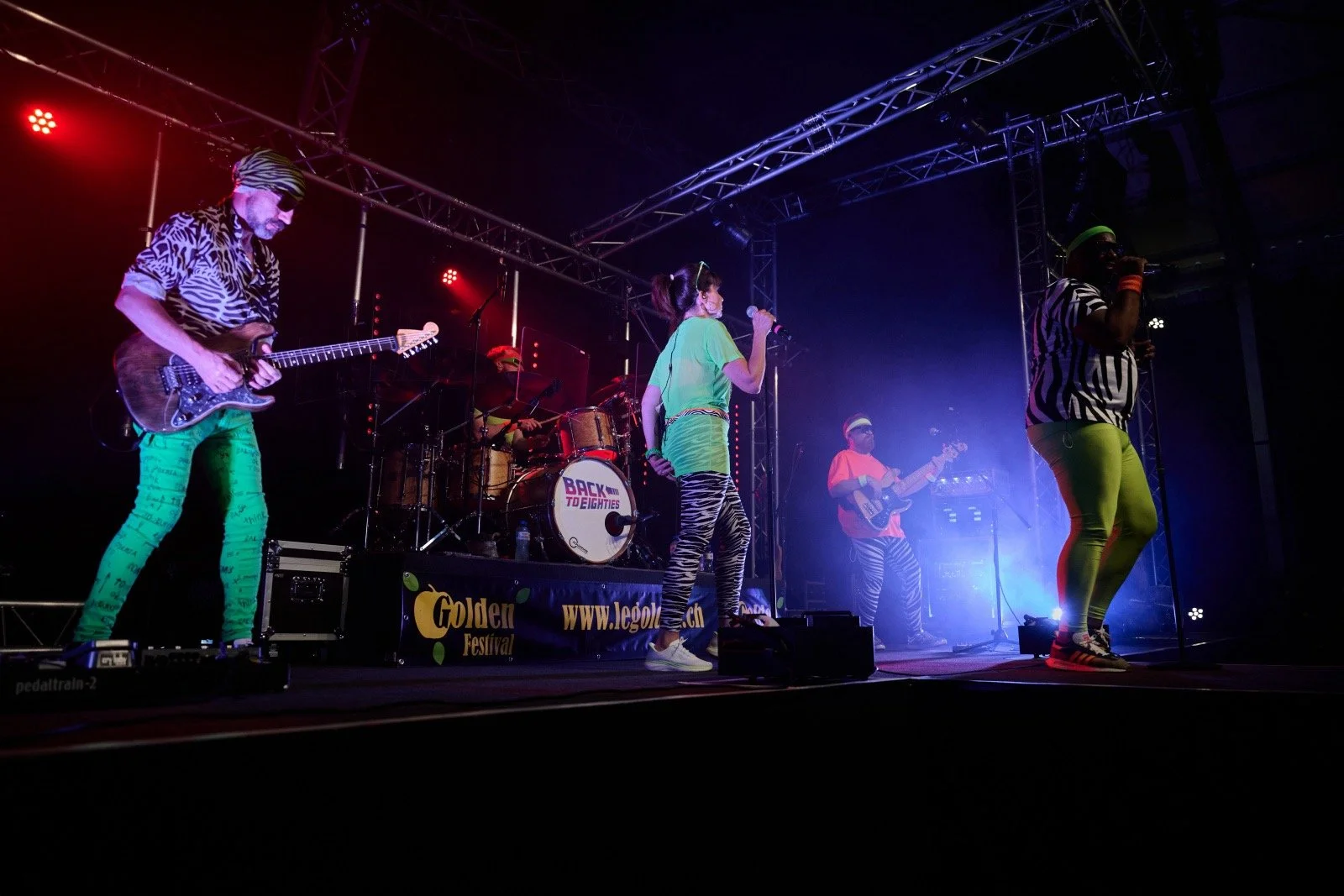 A band of five performers on stage wearing colorful and zebra print clothing, playing instruments and singing during a live concert at the Golden Festival.