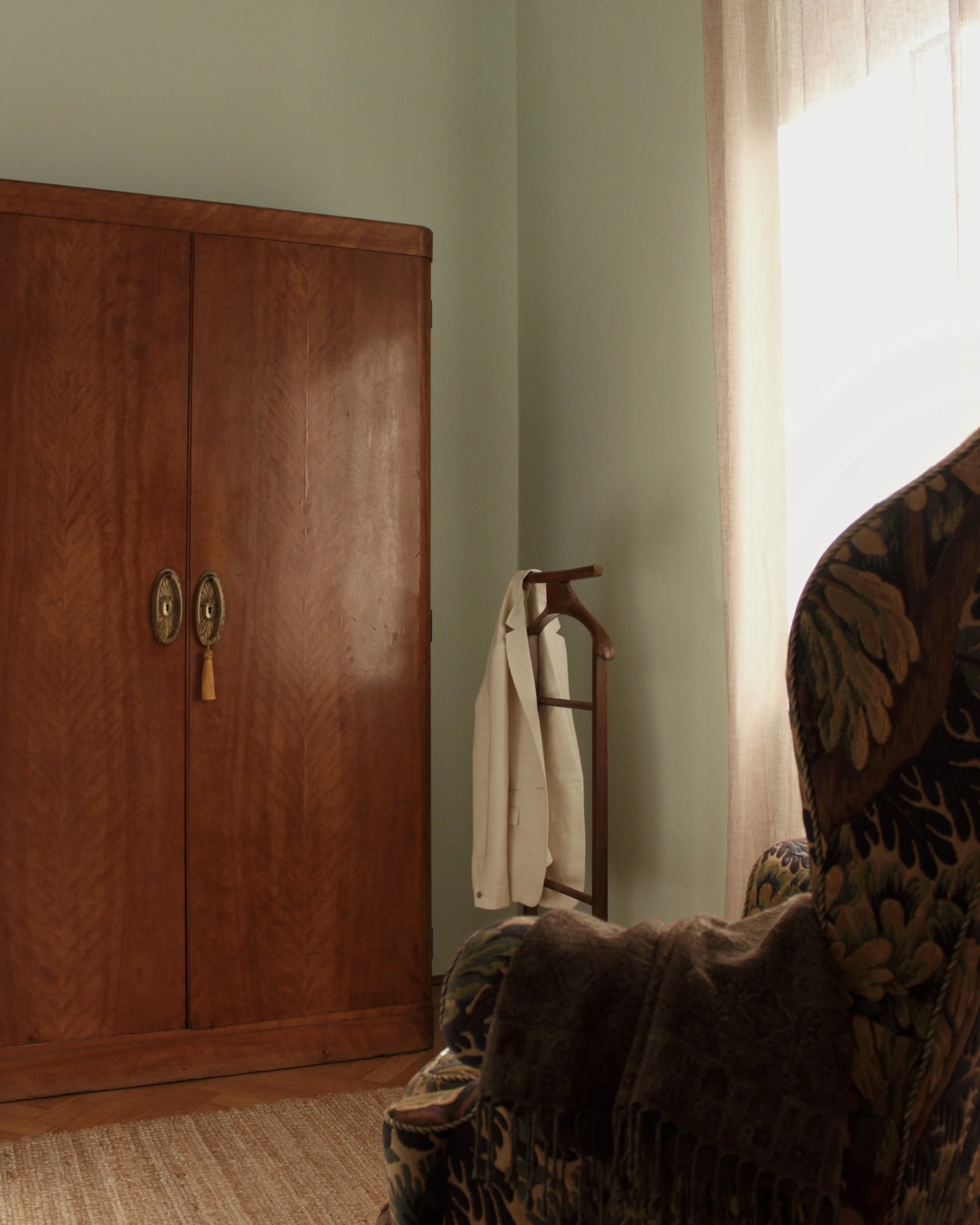 A cozy room with a wooden wardrobe, a floral upholstered armchair with a dark blanket, a wooden clothes rack with a beige coat, and a window with beige curtains letting in natural light.