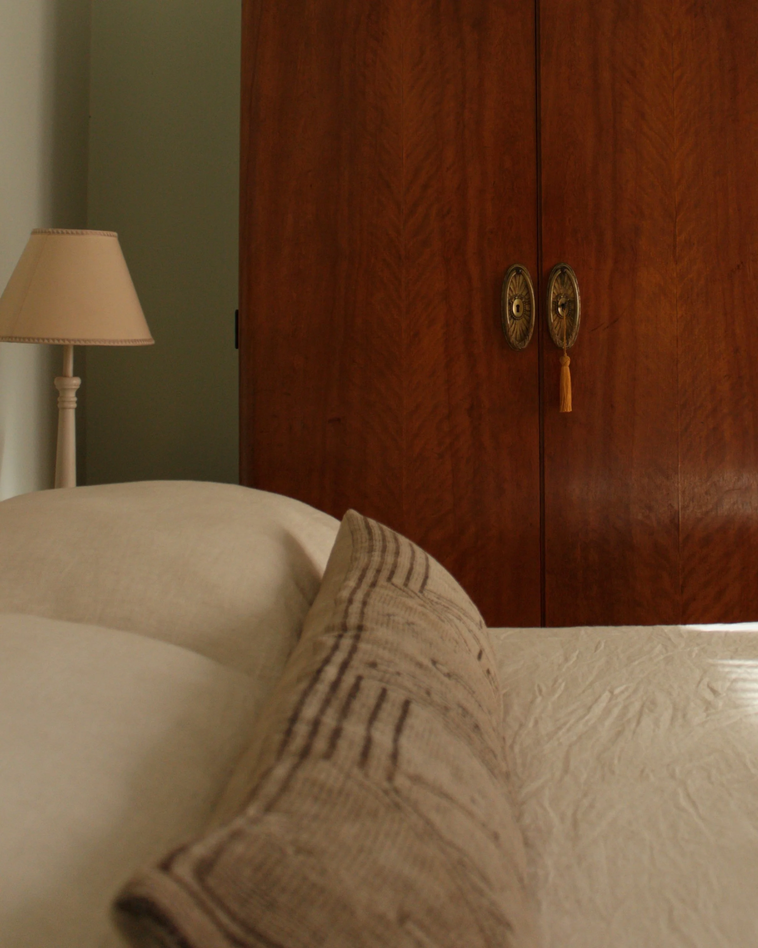 A beige bed with a decorative pillow in front of a wooden wardrobe with round brass handles and tassels. A beige lamp with a shade is on a nightstand to the left.