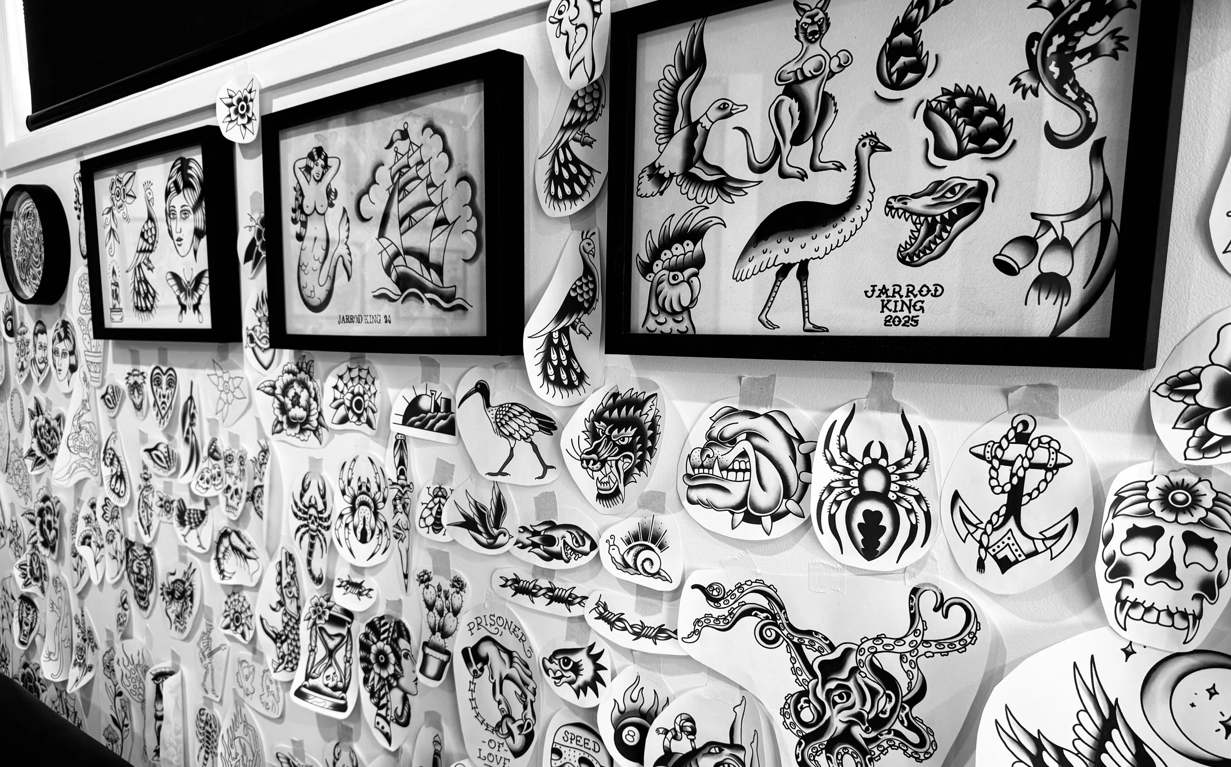 Black and white photo of a wall decorated with numerous traditional tattoo flash designs, including images of animals, skulls, flowers, and various symbols, with framed artwork features of birds, a mermaid, and a sailing ship.