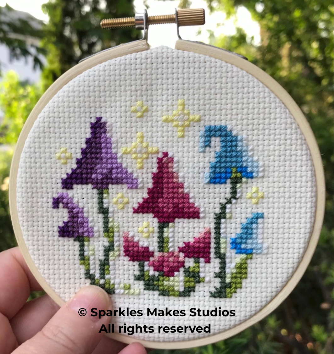 Cross-stitch embroidery of colorful flowers, including purple, pink, and blue, with small yellow accents on white fabric in a hoop
