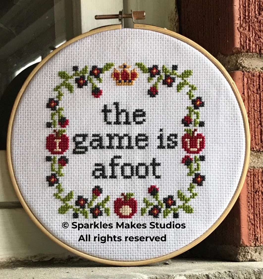 Cross-stitch embroidery hoop with a floral wreath and the words "the game is about" stitched in black thread, with red mushrooms and flowers decorating the design.