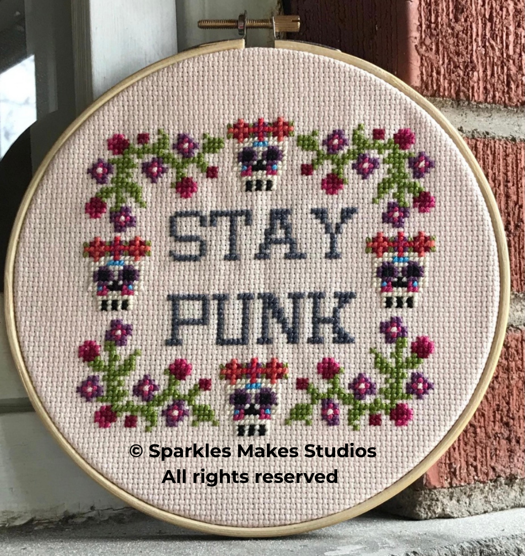 Cross-stitch embroidery in a hoop with the words "STAY PUNK" surrounded by a floral border and skulls. The design includes colorful flowers, skulls, and the phrase in grey stitches on a pink background.