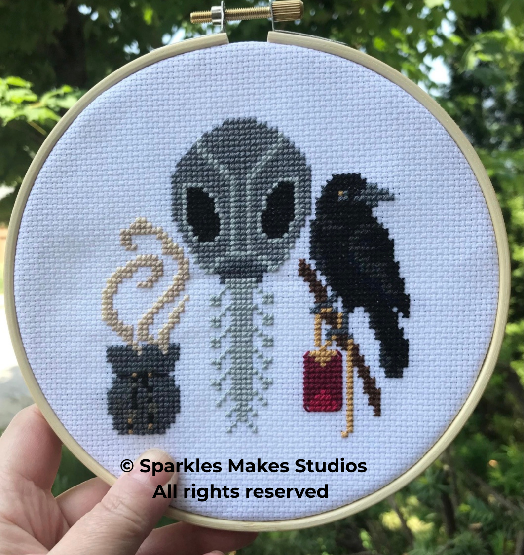 Cross-stitched artwork of an alien with a skeletal spine, holding a smoking cauldron on the left, and a crow with a red book and staff on the right, in an embroidery hoop.