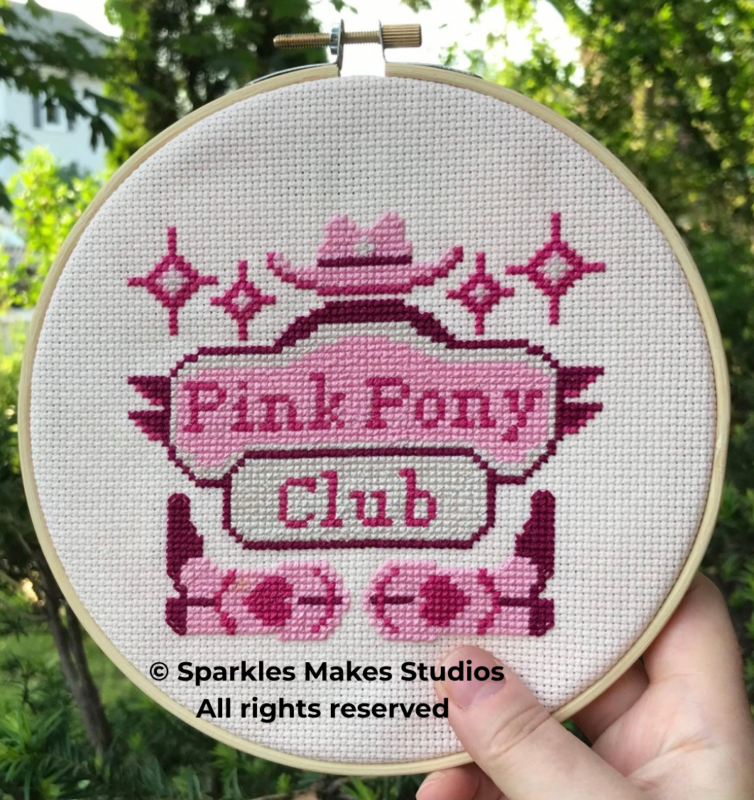 Cross-stitch embroidery of a pink pony themed logo reading 'Pink Pony Club' with a pink hat on top and pink hearts below, in a wooden hoop held outdoors against a background of green trees.