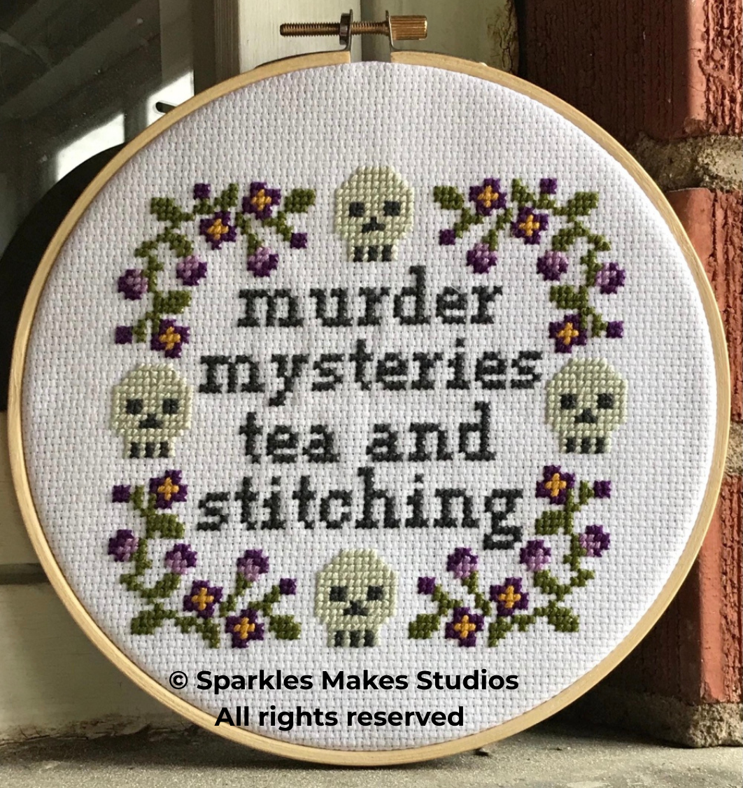 Cross-stitch embroidery with purple flowers, white skulls, and the text 'murder mysteries tea and stitching' inside a wooden hoop.