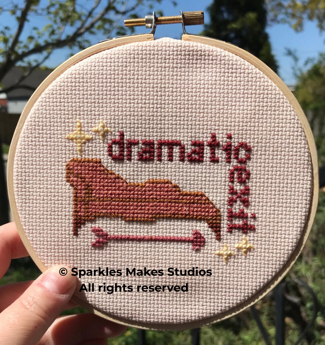 Cross-stitch embroidery hoop with a design of a red and brown mountain with a pink arrow underneath, and the words 'dramatic' and 'rescue' stitched in red and brown respectively, surrounded by small decorative stars, on a pink fabric background.