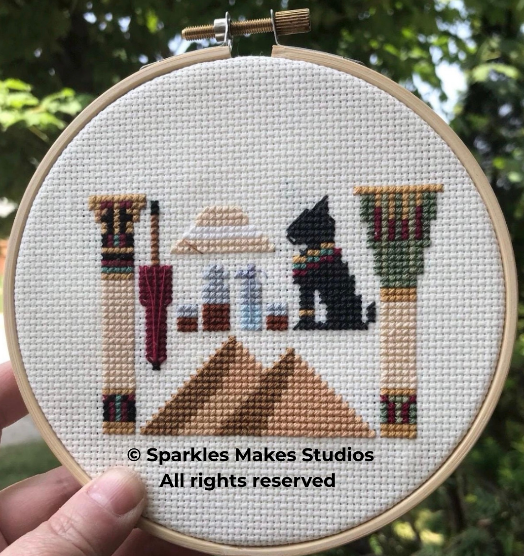 Cross-stitch embroidery of a tabletop scene with an Egyptian cat, pyramids, a sun, and decorative pillars.