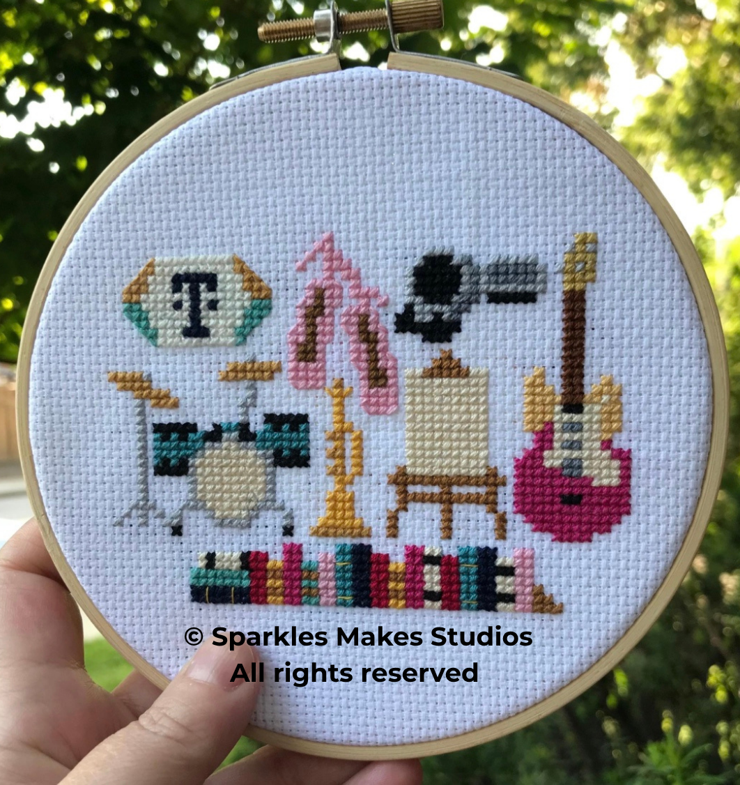 Cross-stitch embroidery featuring musical instruments: a drum set, a trumpet, a microphone on a stand, and an electric guitar; a decorative letter T; a pink scarf; a small wooden table with a canvas on it; a multicolored book cover at the bottom; aga