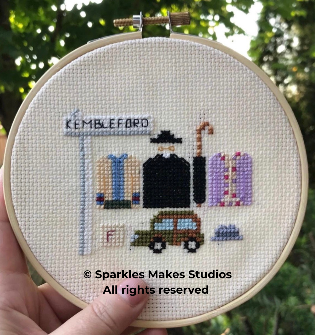 Cross-stitch embroidery depicting a train station scene with a station sign labeled 'KEMBLEFORD', a person dressed in black with a hat and umbrella, a vintage car, a small pile of rocks, and a pink train behind the person. The stitched scene is frame