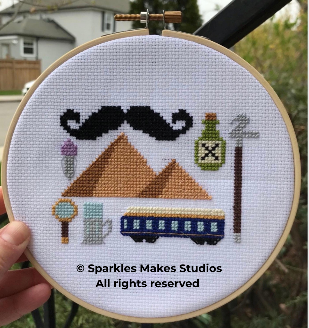 Cross-stitch embroidery of a man's face with a large black mustache, pyramids, a train, an ice cream cone, a pocket watch, a green bottle, and a smoking pipe on a white fabric background in a wooden embroidery hoop.