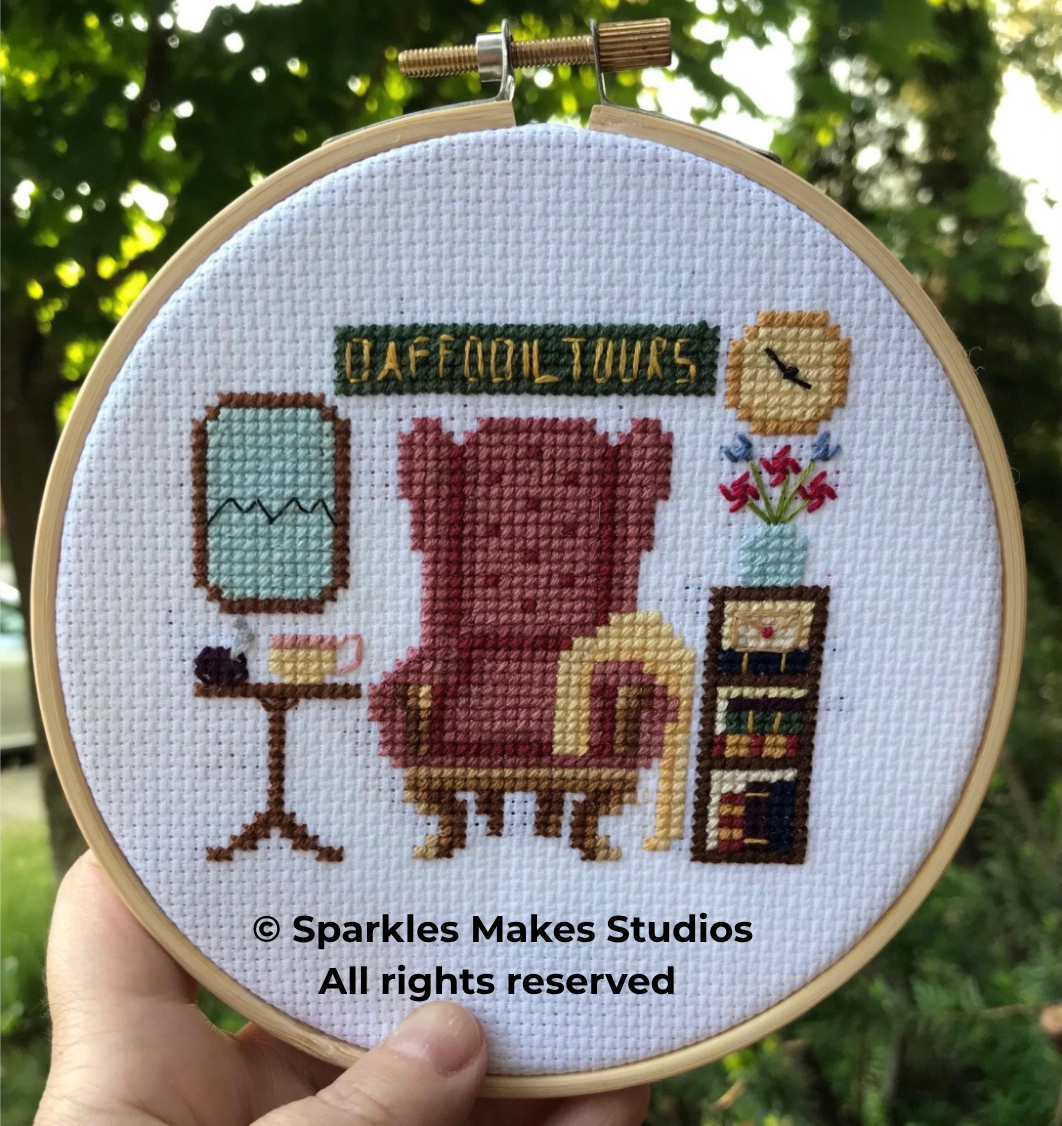 Cross-stitch embroidery of a teddy bear sitting in an armchair in a room with a lamp, a clock, a bookshelf, and a vase with flowers. The words "DAFFODIL TOURS" are stitched in a banner at the top.