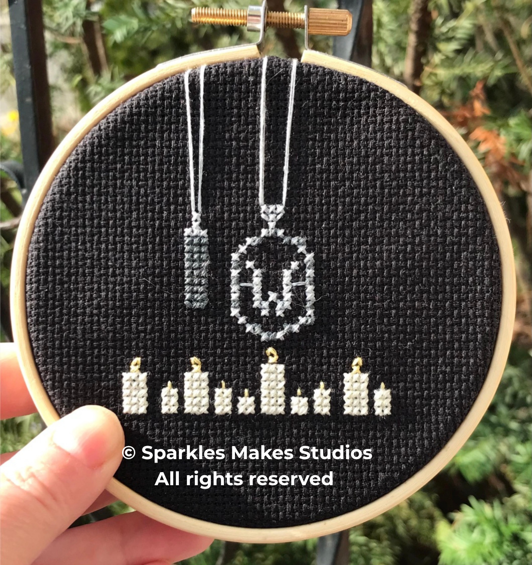 Cross-stitch embroidery of a Halloween-themed design featuring a ghost with candles and hanging dog tags, set on black fabric. The image includes a copyright text overlay from Sparkles Makes Studios.