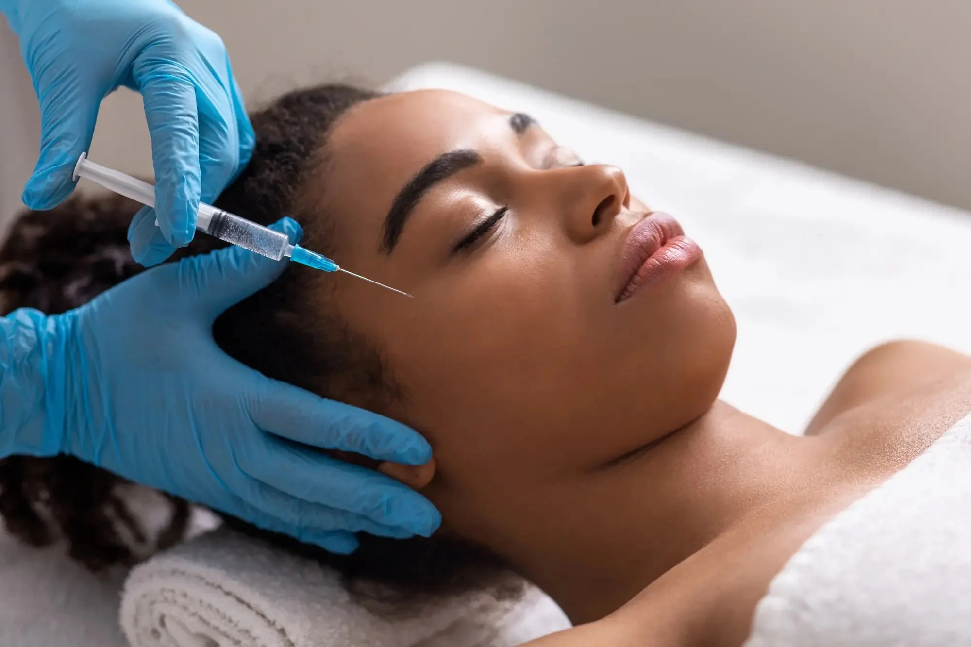 A woman receiving a facial injection treatment while lying down with eyes closed, a healthcare worker wearing blue gloves administering the injection near her temple.