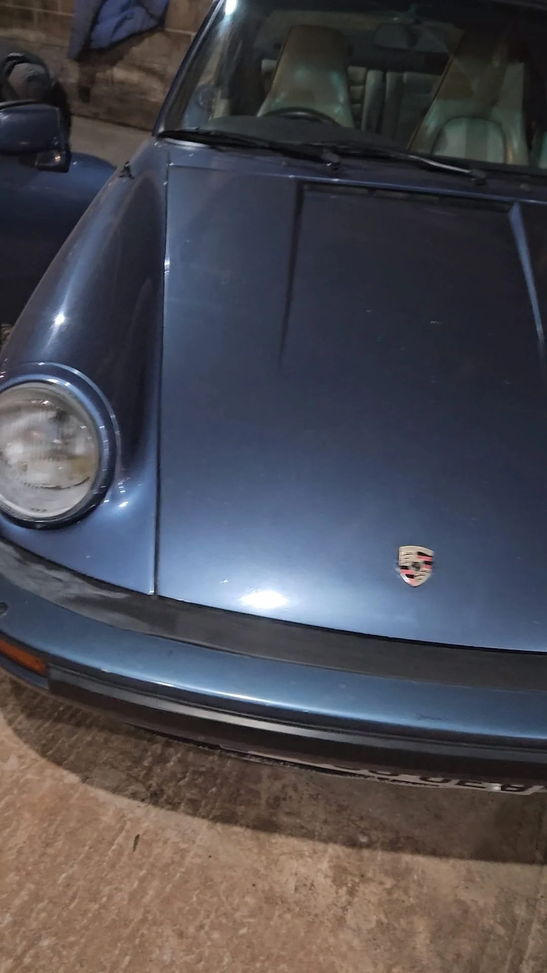 Front view of a vintage blue Porsche sports car with a visible Porsche logo on the hood, parked inside a garage.