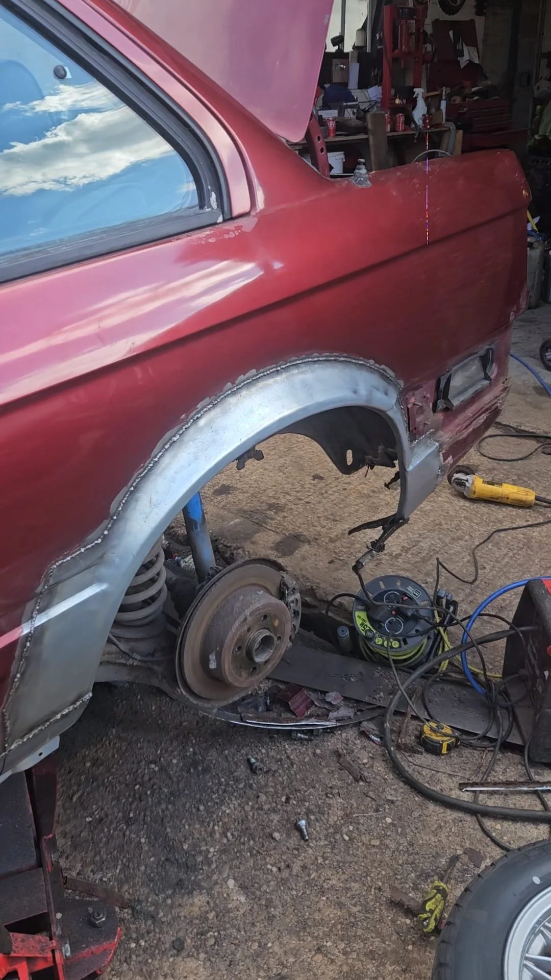 A red classic car is being restored or repaired, with the rear panel and wheel area exposed, showing suspension, brake rotor, and unpainted metalwork, in a garage workshop.