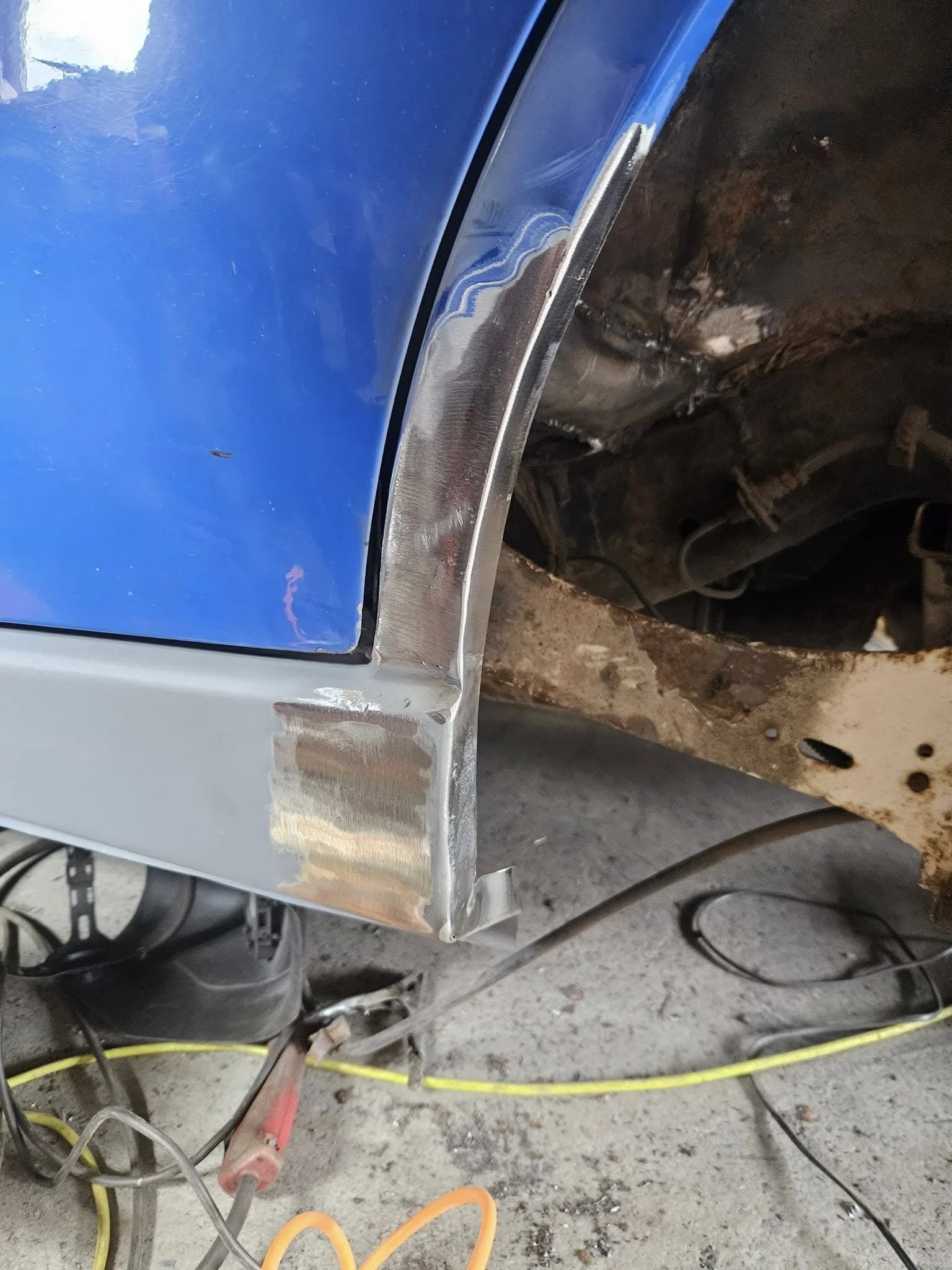 Close-up of a car's front fender with some bodywork and rust repair in progress, showing a blue and silver painted section with exposed rusty metal underneath.