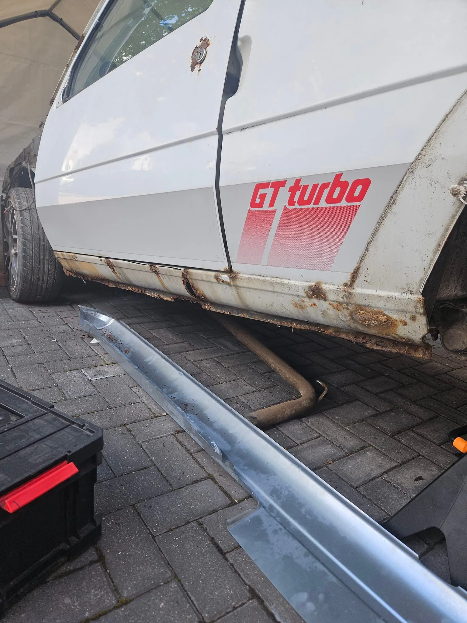 A white car with rust and damage on the bottom side, featuring a red and gray 'GT turbo' decal. The car is missing the front bumper, which is lying on the ground nearby, and appears to be undergoing repair.